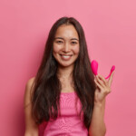 Glad brunette woman poses with smart bluetooth vibrator, uses special app on mobile to improve orgasm, holds sex tool to enhance pleasure, isolated on pink wall. Modern technologies and sexual life